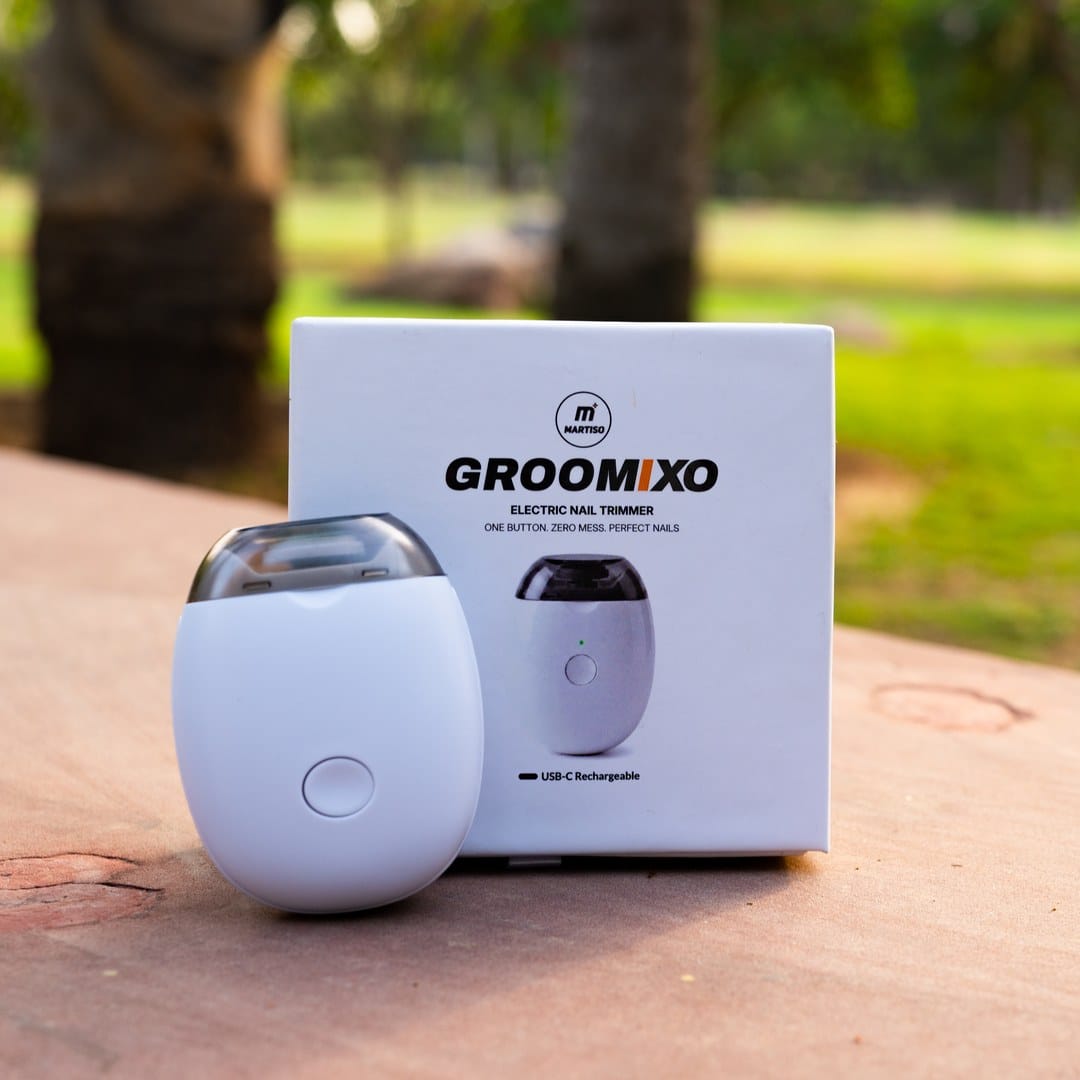 A white Groomxo electric nail trimmer and its retail box displayed on a wooden table with a blurred outdoor garden background.